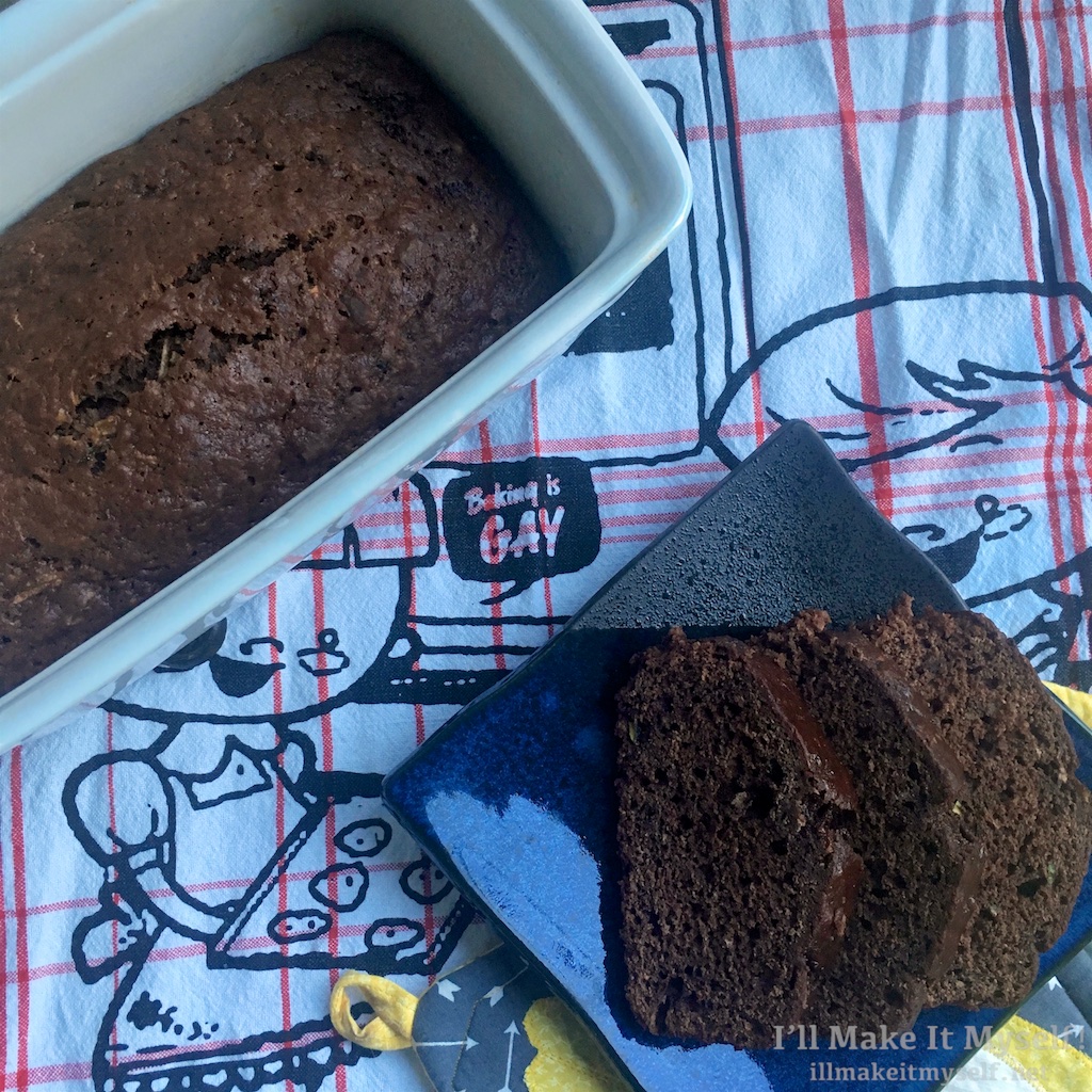 Image of a loaf of chocolate zucchini bread and some slices of the bread on a "Baking is Gay" towel