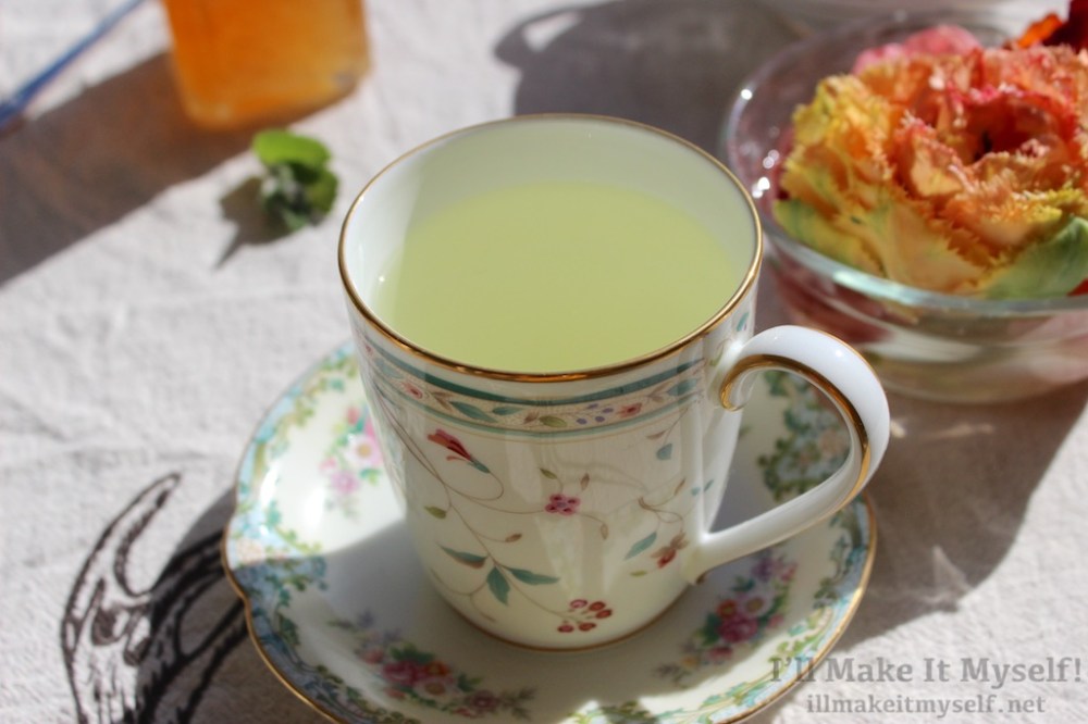 Image of a tea cup with a floral pattern and gold full of mint tea. The cup and saucer are on a table with red and orange rananculus flowers in a bowl.