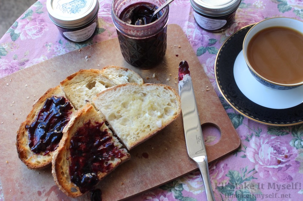 Image: two slices of toast on a cutting board. One half of each toast has purple jam on it and the other half has only butter. There are three jars of jam on the table; one is open and has a spoon in it. There is a cup of coffee in a porcelain cup with a sauce.