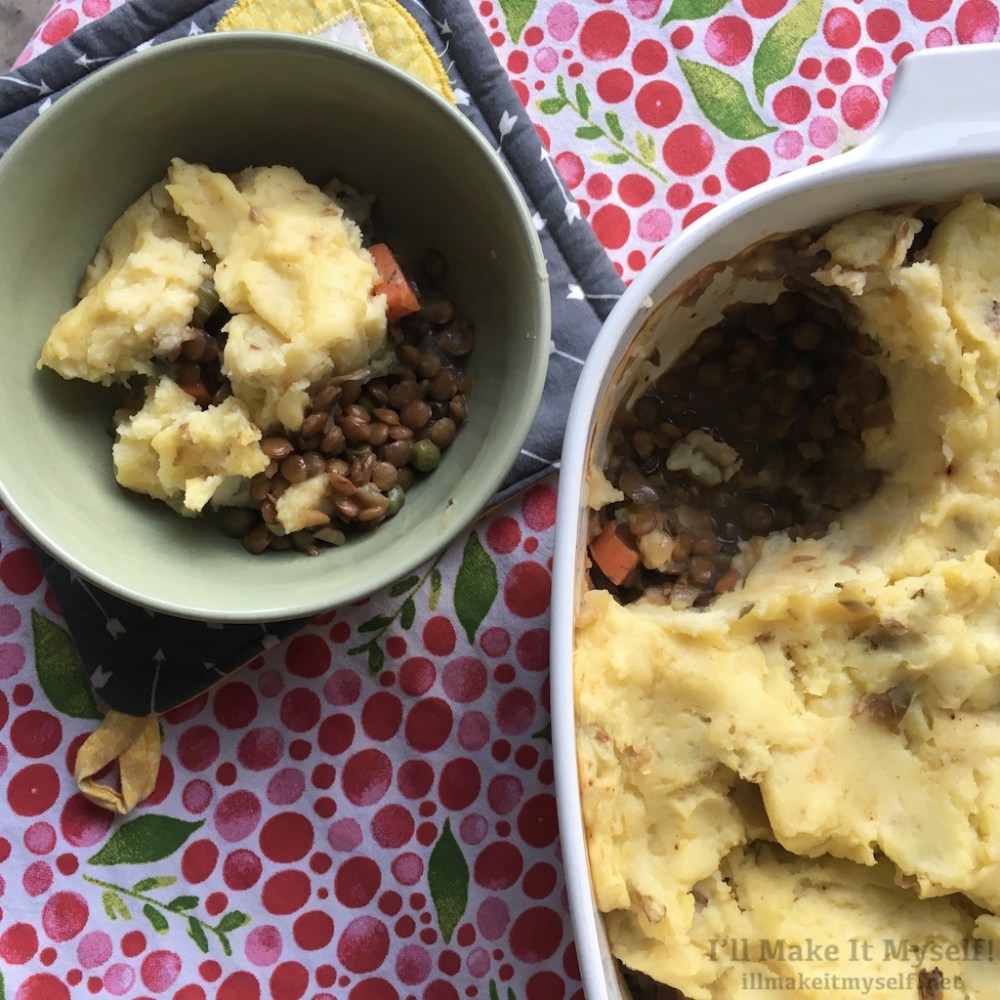 Image: a blue-flower Corningware with shepherd's pie. There is a green bowl with some shepherd's pie scooped into it. The backdrop is a cloth with red and pink berries and green leaves.