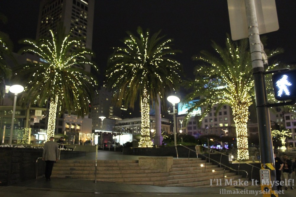 Image: Union Square, San Francisco. The stairs where Johnny walks past on the way to his surprise birthday party. This is a night scene. There are three palm trees with fairy lights on them.