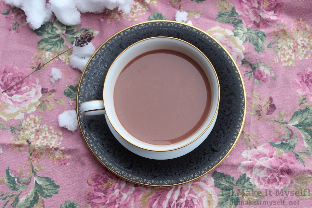 Image: a cup of hot cocoa in a black and gold Noritake tea cup and saucer. The background is a pink floral cloth with snow on it.