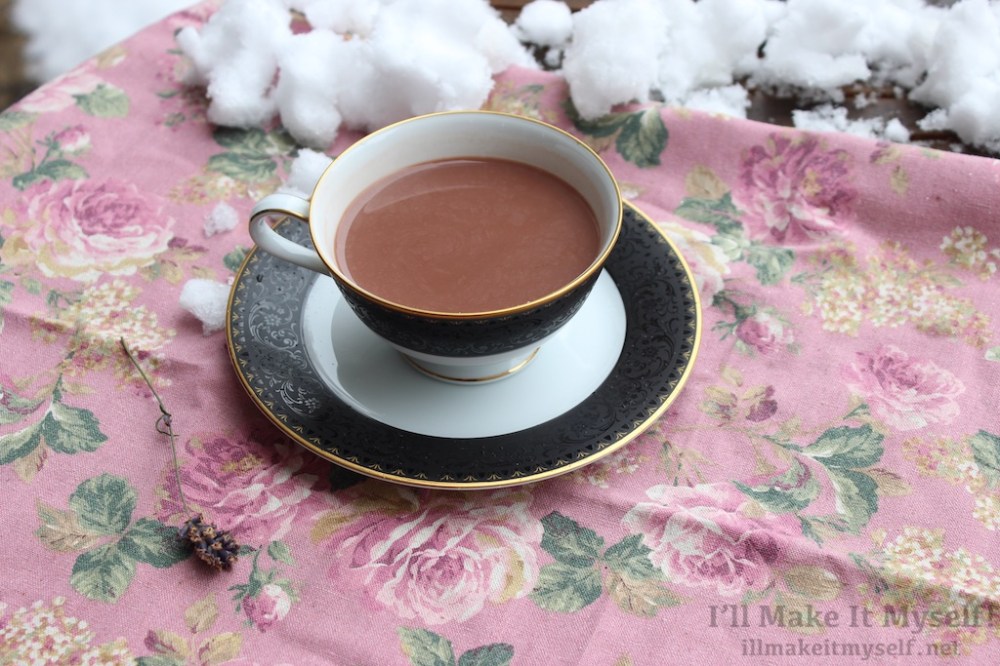 Image: a cup of hot cocoa in a black and gold Noritake tea cup and saucer. The background is a pink floral cloth with snow on it.