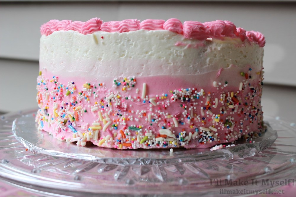 Side view of the cake. The frosting is a white to pink ombré and covered in multicolor sprinkles. 