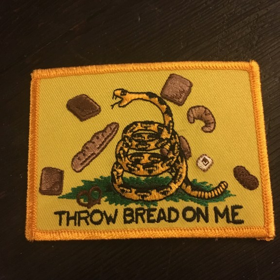 Image: a yellow embroidered patch with a picture of a snake and various types of bread. The text reads Throw Bread on Me, which is a reference to the Revolutionary slogan 