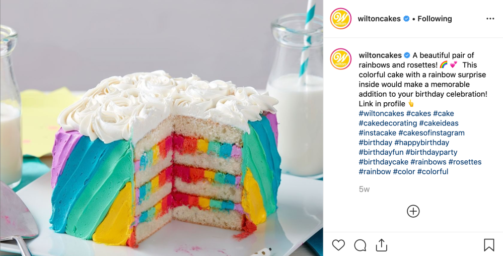 A screenshot of Wilton Cake's Instagram. There is a cake decorated with rainbow frosting on the outside and between the layers and white frosting rosettes on top. The text reads, "A beautiful pair of rainbows and rosettes! 🌈💕 This colorful cake with a rainbow surprise inside would make a memorable addition to your birthday celebration! Link in profile 👆 #wiltoncakes #cakes #cake #cakedecorating #cakeideas #instacake #cakesofinstagram #birthday #happybirthday #birthdayfun #birthdayparty #birthdaycake #rainbows #rosettes #rainbow #color #colorful"
