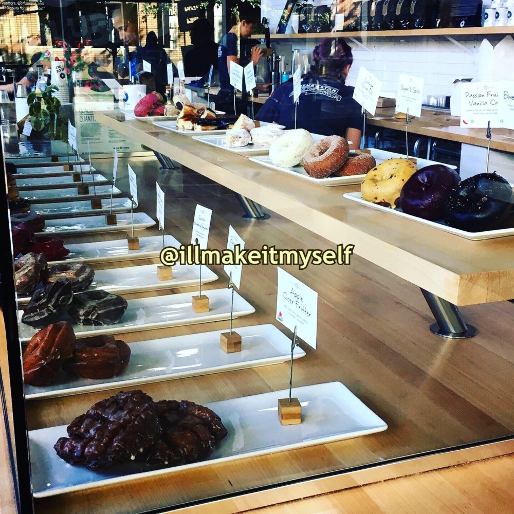 The counter at Blue Star Donuts with all the donughnuts lined up on plates behind glass. There are about 15 varieties. The display is minimalist and clean.