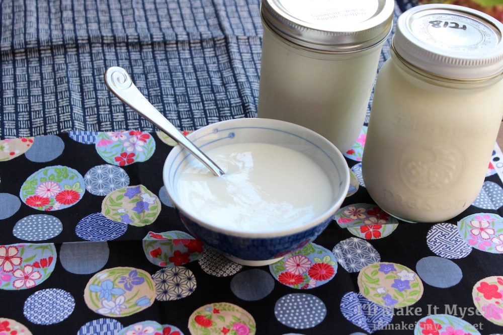 A bowl of yogurt with a spoon in it. There are two pint jars of yogurt next to the bowl. The bowl has a spoon with a spiral pattern. The cloth under the bowl is a Japanese pattern with circles with flower and geometric designs, and the cloth in the background has blue and white cross-hatching.