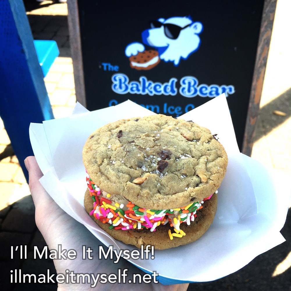 An ice cream sandwich with two peanut-butter chocolate chip cookies, coffee ice cream, and rainbow sprinkles. The sign in the background says The Baked Bear Premium Ice Cream Sandwiches and shows a blue cartoon bear with sunglasses eating an ice cream sandwich.