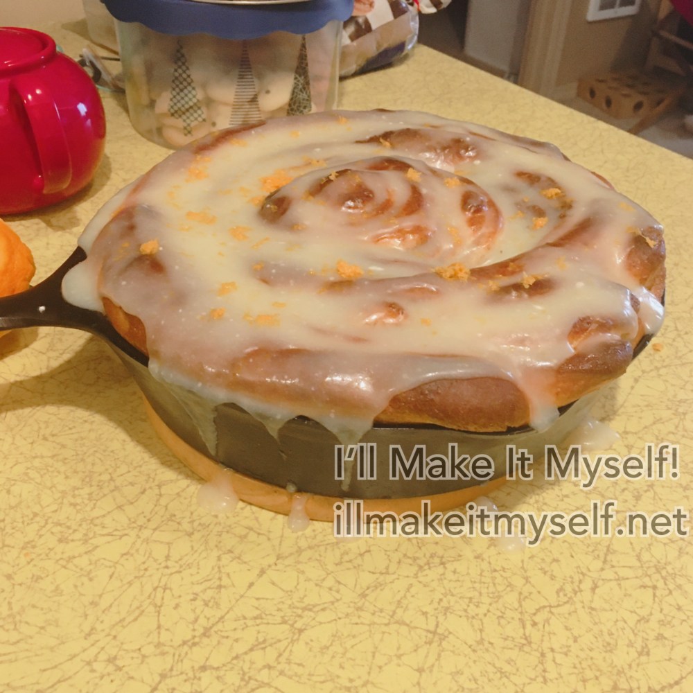 A huge cinnamon roll topped with too much frosting and some orange zest, on top of the yellow formica table. Tea pot for scale.