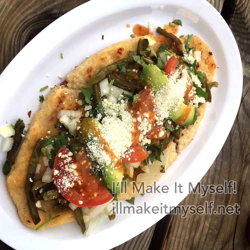 Tlacoyos on a paper plate. The tortilla is stuffed with beans and topped with cilantro, cheese, vegetables, and tomatoes.