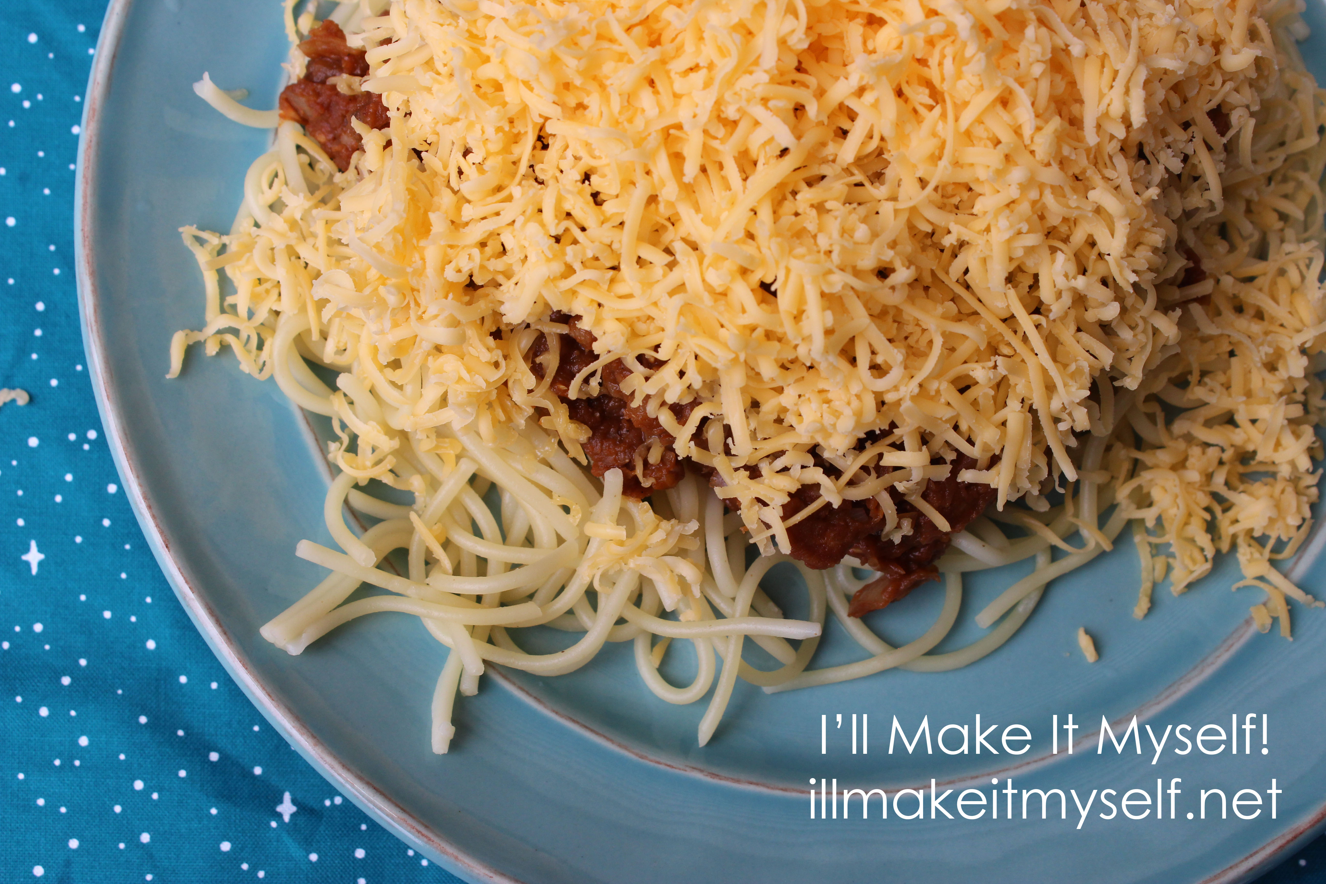 A plate of vegetarian Cincinnati chili with red lentil chili sauce. The chili is on top of spaghetti and covered with a mound of cheddar cheese. The plate is on a blue cloth with a star pattern from Fireside Textiles.