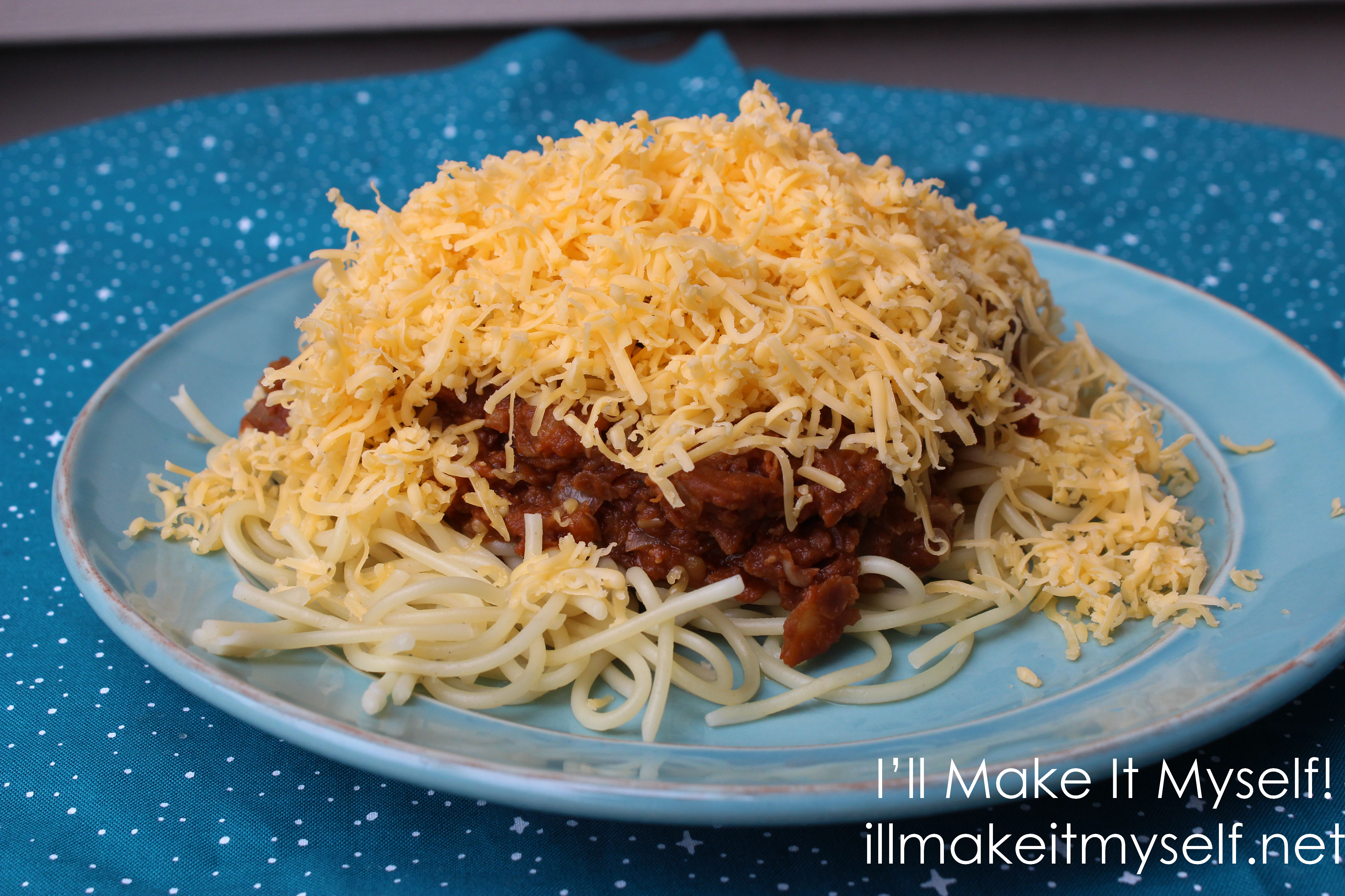 A plate of vegetarian Cincinnati chili with red lentil chili sauce. The chili is on top of spaghetti and covered with a mound of cheddar cheese. The plate is on a blue cloth with a star pattern from Fireside Textiles.