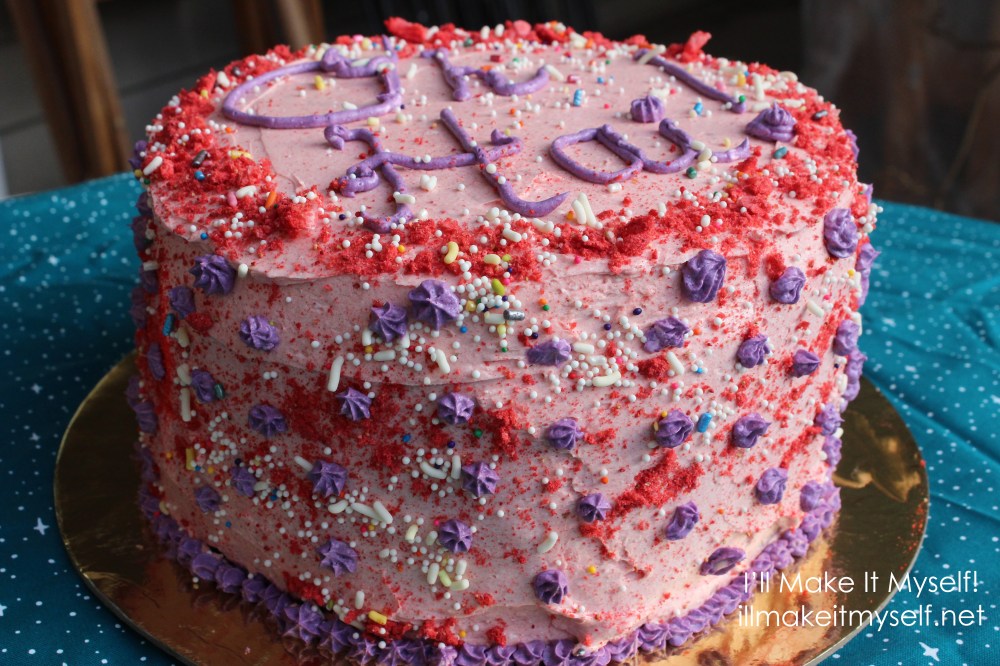 Side view of the cake. A three-layer cake frosted in pink buttercream. There is a dusting of crushed freeze-dried strawberries around the edge of the top layer. The cake is covered in rainbow ball sprinkles and jimmies. There is text on the cake in purple icing: "Oh Hai!" There are also purple stars piped on the sides and the edge of the bottom layer. The cake is on a gold cake round on a blue cloth with white stars.