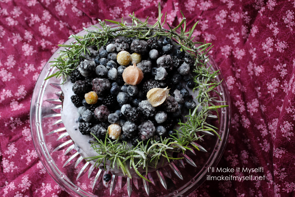 Goblin Market Cake: a two-layer chocolate cake on a crystal cake stand. The cake is frosted in a light-purple coconut whipped cream and topped with sugared rosemary in a semi-circle and sugared blackberries, blueberries, and yellow ground cherries. Overhead view.