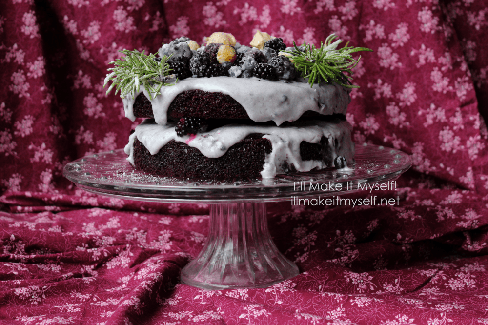 Goblin Market Cake: a two-layer chocolate cake on a crystal cake stand. The cake is frosted in a light-purple coconut whipped cream and topped with sugared rosemary in a semi-circle and sugared blackberries, blueberries, and yellow ground cherries. Side angle.