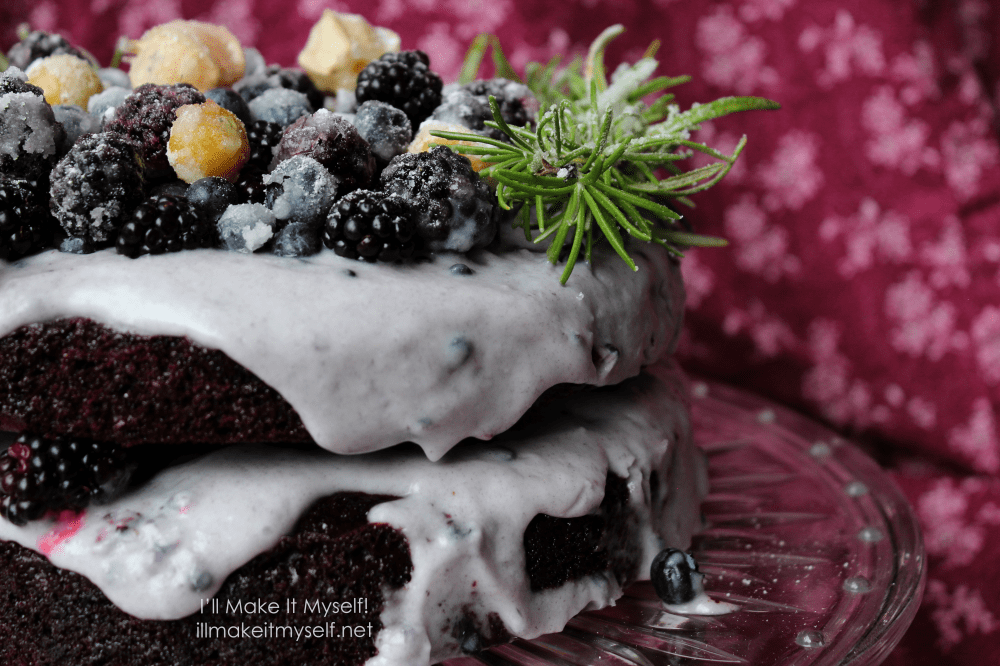 Goblin Market Cake: a two-layer chocolate cake on a crystal cake stand. The cake is frosted in a light-purple coconut whipped cream and topped with sugared rosemary in a semi-circle and sugared blackberries, blueberries, and yellow ground cherries. Detail of the side of the cake; one blueberry has fallen off.
