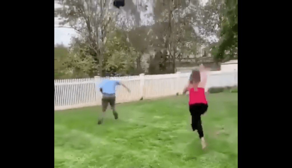 The father-to-be and a party-goer chase after the escaped black balloon towards a white picket fence.