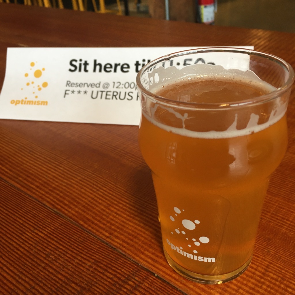 Photo of a small glass of beer in an Optimism Brewing glass in front of a placard that reads "Sit here till 11:50 am. Reserved at 12 pm for F*** Uterus Party."