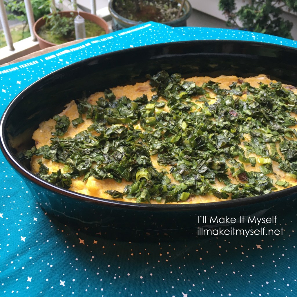 Polenta casserole topped with sauteed green spring onion stems. The casserole is in an oval black casserole dish and is on top of a piece of turquoise fabric with a starry sky pattern.