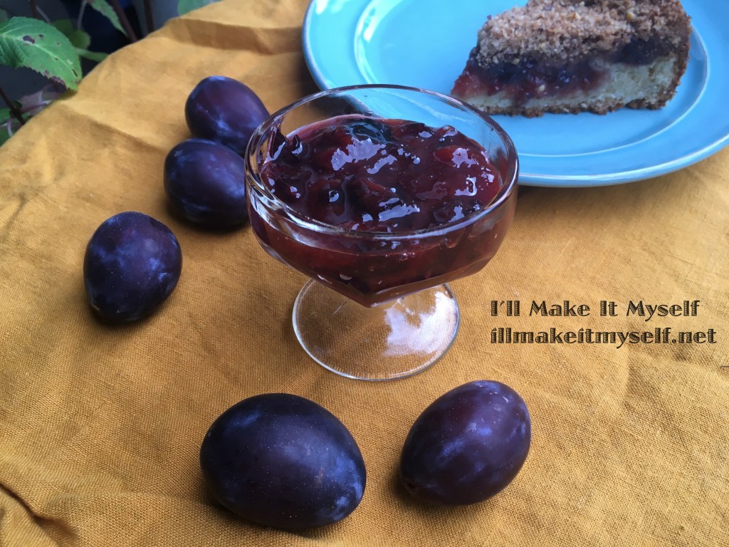 Photograph of a glass dish full of plum-cardamom jam. The dish is set on a mustard-colored linen cloth with a few Italian plums placed around the dish. In the background is a slice of coffee cake with plum jam filling and topped with streusel. 