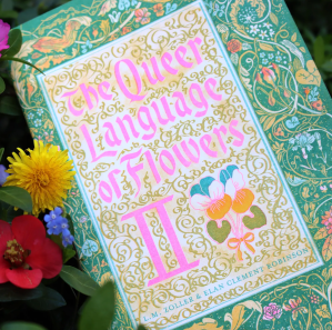 Photo of the cover of The Queer Language of Flowers Volume 2 zine. The risograph-printed cover has the text in pink, an illustration of a pansy in yellow and blue, and a decorative floral pattern with a green background.